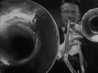 A clip from a black and white silent film, including quick cuts between various social dances. The quick cuts include two pairs of dancers in a ring, a band playing showing various musicians and close-ups on instruments, and couples dancing in the center of a restaurant-like setting, with other guests watching. The clip ends with a close-up on the dancers’ feet.
