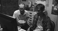 A black-­and-­white shot from The Great Buddha+ showing two ragged men watching video footage on a computer screen in a small room. One holds a plastic bowl of rice.