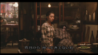 Image of someone standing and facing someone who is sitting at a table at a restaurant. Both are wearing plaid. In the deep background, there are red and pink banners with black calligraphy on them. There is also white calligraphy in subtitles superimposed on the image.