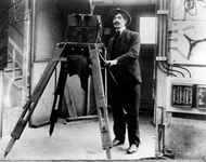 Photograph of a man in a suit standing by a large, boxy apparatus on a large tripod.
