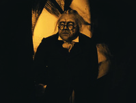Fearsome Dr. Caligari, center, looks with inquisitive menace off Left in his deep black suit and shining gold lapel and tie. The frame is gold tinted. Behind him center frame (for the Right and Left are simply black) is a bright, indescribable, non-­representational background, partially hand-­drawn, partially painted with black streaks that simulate shadows. The background vaguely resembles a large and small flower petal or parabola of light.