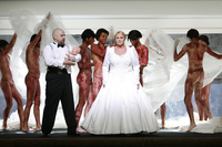 A man in a tuxedo and woman in a wedding dress are surrounded by seven naked men covered in blood. He holds a baby doll, also smeared with blood.