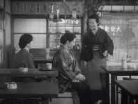 Two women sit and one stands, smiling, in a restaurant. They are the only people in view and there are empty tables behind and in front of them. Menu items for this restaurant are tacked on the walls, and a calligraphic noren in white is visible across the street.