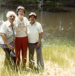 Each wears a short-sleeved shirt and long pants. They stand in tall grasses with a pond and a glimpse of woods behind them. All smile toward Cohen, who is taking the picture.