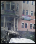 This building is the courthouse, located along the main street running through the city of Khorog. This was one of the buildings that was burned down by protesters during the conflict on May 21, 2014. The picture shows burned-out windows of a two-story building and a car that is charred and destroyed. Rubble surrounds the buildings and car.
