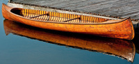 A color photograph of a beautiful cedar strip canoe built by the Lakefield Canoe Company of Peterborough ca. 1925–1930. The canoe uses three wide boards as planking on either side. It is pictured in the water, next to a wooden dock.