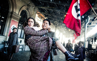 Pu Jizhou carrying the pregnant Rosa as they rush to catch the last train leaving Austria. It is snowing, and Nazi flags are prominently displayed throughout the train station. Both look anxious and exhausted.