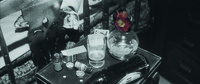 Close-­up, in lower center frame, of a small end table. An overturned beer bottle, some medicine bottles and small boxes, two white pills and a glass of water scattered about the table. Also on the table, stemless vermillion camellia, its hand-­painted color contrasting with the monochrome frame, sticks out of a tiny glass vase.