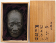 A wooden box, opened to show a brownish-black mask of a human face set in purple cloth, alongside the lid which has black calligraphic text painted on it.
