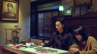 A shot from The Bold, the Corrupt, and the Beautiful, showing a mother holding a brush painting of flowers in a Japanese-­style living room. Her bored daughter watches at her side.