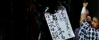 Black calligraphy is written on white paper sign at a festival. Black calligraphy forms a decorative pattern on a white kimono worn by a man.