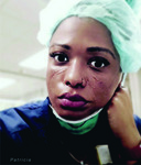 In a tight close-up, a middle-aged Black woman with a small golden nose ring and expressive eyes gazes solemnly at the camera, her chin resting on her left hand. She wears a protective hair net. Her face is scarred and tear-stained.