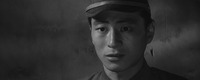 Close-­up of Mikami in his uniform and cap, off-­center Right, his face full of dismay, neurotic apprehension; emphasis also on the blank stuccoed wall stretched out behind his head.
