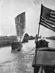 A black-and-white photo showing a single-sail boat on a canal, with a few more single-sail boats in the background. A large American flag hangs on a flagpole from another boat.