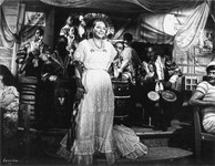 A black and white photograph of Toña la Negra in an all-­white version of traditional Veracruzan costume in En carne viva. She is on stage surrounded by musicians.