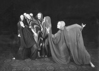Photograph of “Dance of Death II,” in which the tormenter beast lunges toward Wigman, the tormented martyr, who is supported by six group dancers atop carpet floor and in front of black background. Everyone wears billowing sheer fabric outfits with veils and hard masks. Wigman wears a more narrow dress than the others with dark fabric and lighter color chevron stripes going down the entire garment. She leans back against the group dancers.