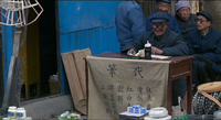 A man sits at a small table, with several people behind him, in a cluttered street. The table has a front draping with calligraphy.