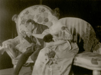 Khlestakov (Garin) and Anna Andreevna (Raikh) sit on an oversized sofa. In this photograph, he holds her pinkie on a teaspoon to kiss it.