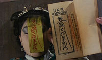 A woman has a streamer with red calligraphy attached to her forehead next to an open pamphlet with black and red calligraphy.