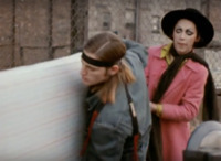 Outdoor urban scene with chain link fence and dark brick buildings with windows. Man carries mattress while hat-wearing trans woman helps.