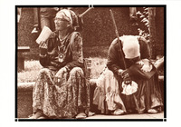 Two women rest, dressed in casual layers of native dress, jewelry, scarves, and moccasins. One holds a pamphlet. The other is slumped over, head down, holding a walking stick.
