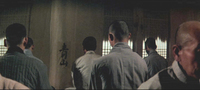 A group of men sit in front of a poster on the wall with black calligraphy printed on it.