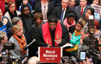 Image of a reverend speaking at a podium, surrounded by press and listeners.