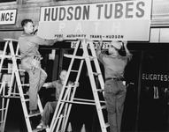 Three men installing a sign that said “Hudson Tubes –­ PATH –­ Port Authority Trans-­Hudson.”
