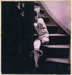 The hand-tinted black and white photograph from The Games of the Doll series (1938-1949) by Hans Bellmer is an indoor shot featuring an armless doll with an amputated leg, tied to a stairway and gazing blankly at the viewer. The doll’s body consists of a hand, placed between the balusters, a head with wig and bow, two pelvises, an abdominal sphere, and two legs, one truncated. The complete leg, the left one, is bound at the knee with twine in the form of a bow, while the other is cut off midway across the thigh. The doll’s body is woven into the balusters, draped on the stairway, hiding its face, while looming large for the viewer.