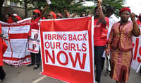 Two women raise their fists and chant. The top of the “#BringBackOurGirlsNow” hashtag can be seen on the front of one of their red outfits, and police officers are visible in the background.