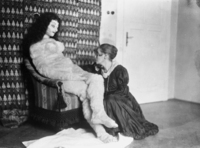 The photograph, presumably taken by her sister Henriette, shows the crouching doll maker Hermine Moos clad in a black dress with a white collar, and the unclothed furry doll, seated in an armchair with her legs crossed. The doll looks like a queen on a throne, granting an audience to a loyal subject. Moos presents herself as a supplicant, kneeling adoringly beside her doll and gazing at her intently.