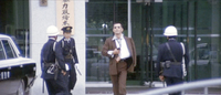 A man leaves a building, surrounded by police. Calligraphy is visible on the signs behind him.