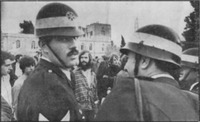 Two policemen photographed from behind with one looking back to the camera. A demonstrator can be seen in the distance between the two.
