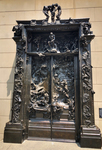 Photo of a bronze cast of the sculpture titled The Gates of Hell by Auguste Rodin. It depicts a doorway with closed doors and figures of demons and humans embedded in the bronze. It stands 19.7 x 13.1 x 3.3 feet and contains 180 figures that range in size from 6 inches high to more than 3 feet high