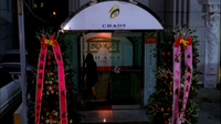 A club has black English text on its sign board and printed on the wall in its lobby. The door is flanked by bouquets of flowers with streamers with black calligraphy printed on them.