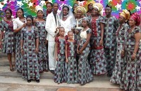 Family members dressed in aso ebi pose for photos around bride and groom against a patterned background.