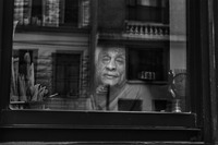 In this black and white photograph, we see the image of an elderly person’s wrinkled face peering out of a window. The subject apparently sits at a desk, as we can see some letters and papers. The face, framed by the window pane, is in soft focus. Reflected in the window we see other buildings and other windows.