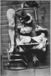 The black and white photograph from The Doll (1934) by Hans Bellmer depicts the doll broken down into body parts ready for reassembly. Laid out on the blueprint are a plastered open torso, a full plastered leg, a wooden hand, a plastered face mask, two glass eyes, a disc with three panoramas, and a wig, framed by two prosthetic arms at the lower corner and a bent prosthetic leg at the upper corner.