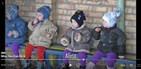 Four young children, all wearing knitted hats and thick winter coats, sit on a bench against a brick wall eating cookies.