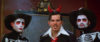Two men dressed as skeletons stand on each side of a man in a suit wearing sequined red devil horns on his head and a coordinating necktie.