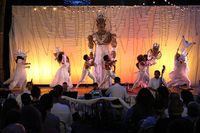 An audience views an orchestra and onstage a gigantic puppet of a fantasy character wearing a crown and white pants. Six actors stand who are half the size of the puppet.