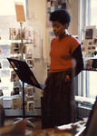 Color photo. McCluskey stands with hands in her long skirt pockets reading her selections from a music stand. Storefront windows and postcard displays are in the background.