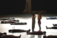 In a large performance space, two dancers hold a balloon in place with their mouths. On the cement floor around them, several performers lie on their backs in a scattered arrangement.