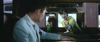 Carefully framed image of Tetsu (foreground, off-­center) in his powder blue suit, seated by the train car window, with Chiharu (middle ground, off-­center Right) in a yellow pantsuit reaching out towards him from the window of another train on the opposite track with the windows imperfectly aligned. Tetsu, head down, refuses to acknowledge her.
