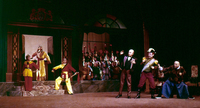 In a Western-style house, six performers pose in two opposing groups. The three on the right appear frightened by the three on the left, who hold swords. A crowd stands outside raising red scarves in the air.
