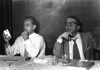 Photograph of Ludwig and Weisenborn seated at a table at the head of a Mittwochgespräch, Ludwig holding an alarm clock.