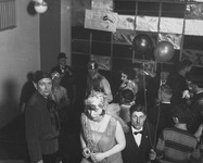 Sepia photograph depicting the staircase from its top at the Bauhaus Dessau during the Metallic Festival. The windows are lined with metal sheets, and the partygoers are dressed in metallic costumes. One of the suited men holds two balloons and stares directly at the camera. Most of the other people are focused on socializing or using the stairs.