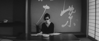 A woman sits at a low table, in front of white calligraphy on the wall.
