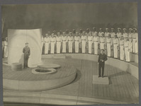 Two characters talk downstage at a distance from one another on a set constructed around simple circles and curves. Upstage of them sailors stand in rows that emphasize the sweeping spiral of the stage.