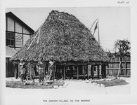 A view of a recreated Samoan house on the Columbian Exposition grounds with Samoan Indigenous people in traditional dress standing or seated before it.
